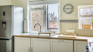Una cucina con frigorifero in acciaio inossidabile, una finestra vista edifici, una sveglia a parete e un cesto sul davanzale.