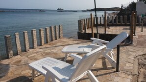 Uma área de estar à beira-mar com cadeiras de plástico branco e um guarda-sol verde, com vista para um mar calmo, afloramentos rochosos e uma ilha distante.