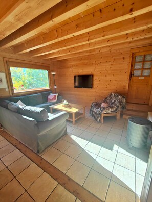 A wooden cabin interior with a sofa, a table, a television, and a window with a view of greenery. - Authentic, cosy chalet for an ideal family holiday (Briançon)