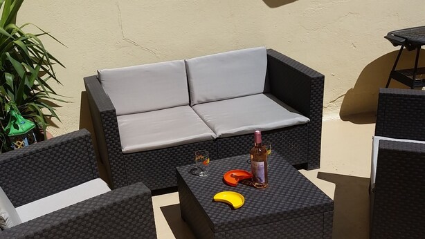 En terrasse med en sort fletsofa, matchende stole og et lille bord med en flaske og glas.
