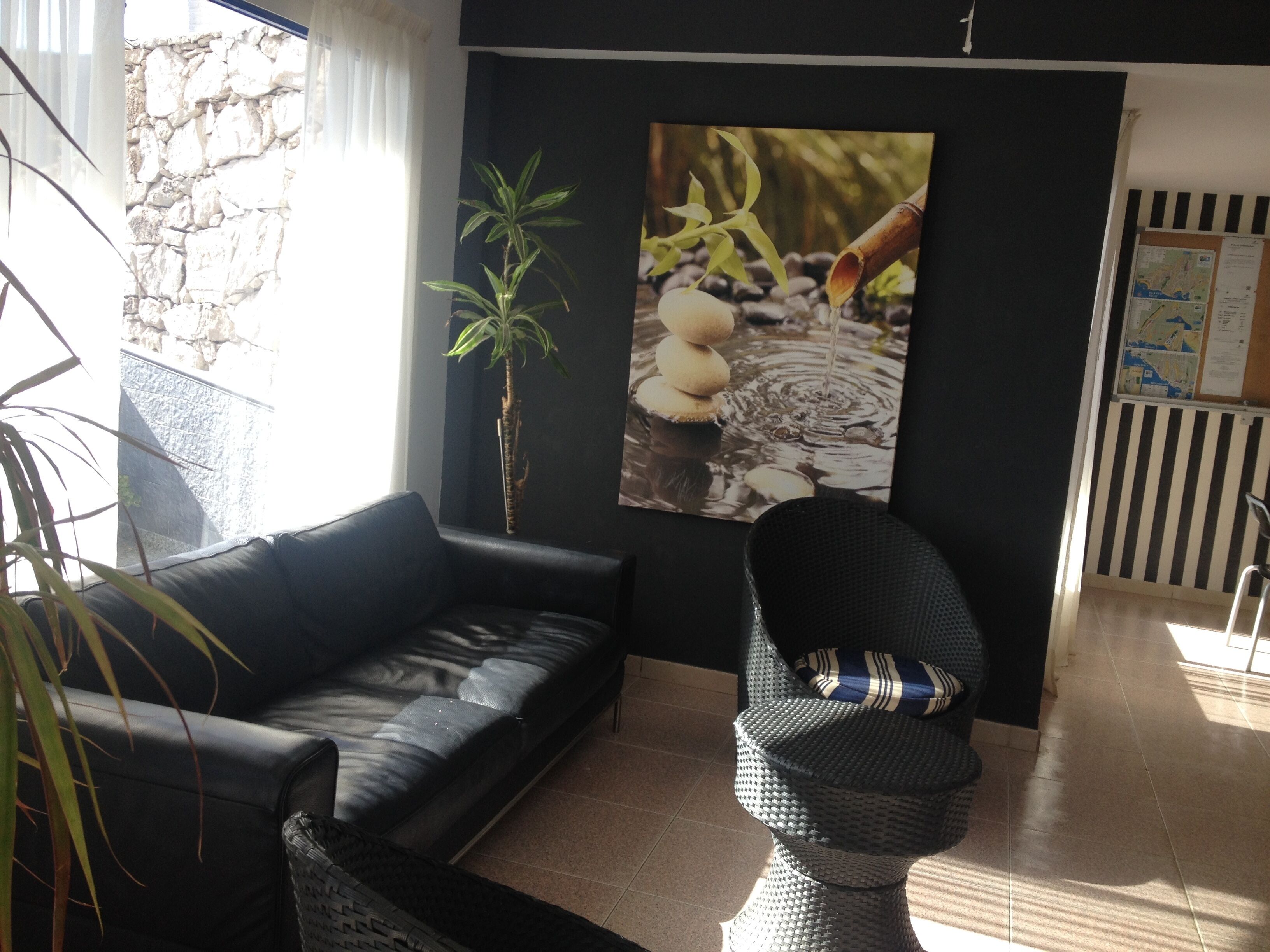 Een woonkamer met een zwarte leren bank, een geweven fauteuil, een groot wanddecoratie van een bamboe waterpartij en een stenen muur.