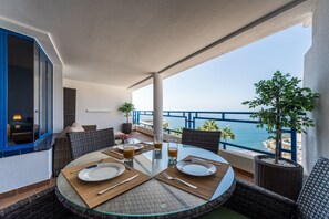 Une table ronde en verre, mise pour deux personnes, avec assiettes, couverts et verres, sur un balcon donnant sur la mer.