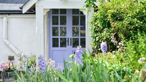 A white house with a blue door, surrounded by lush greenery and purple flowers.