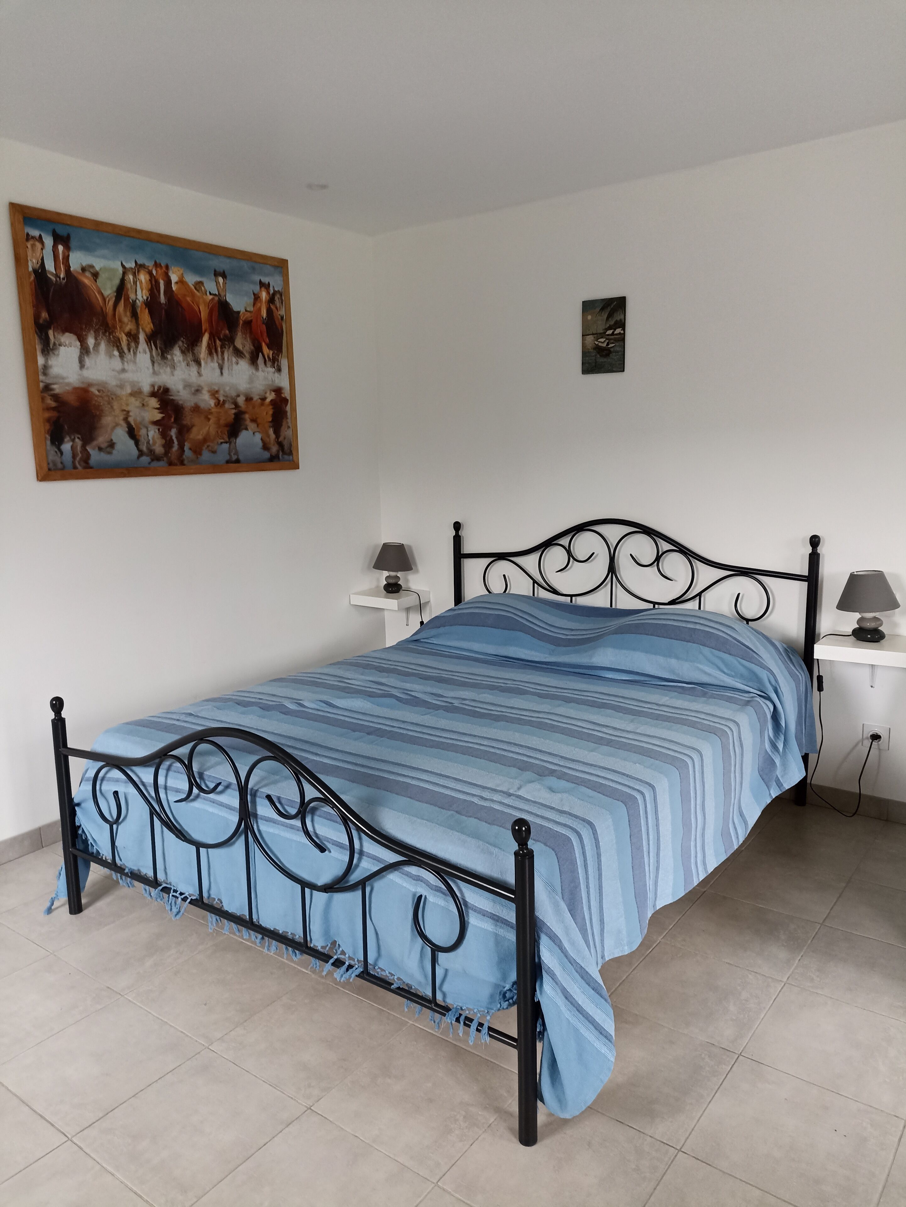 Un lit avec une couvre-lit à rayures bleues, un cadre en métal noir, deux tables de chevet avec des lampes et un tableau représentant des chevaux accroché au mur.