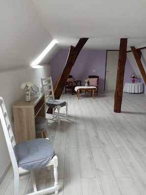 A room with a sloped ceiling, wooden beams, a white chair, a small table, and a door. - cozy cottage ideal for quiet family holiday (Moyaux)