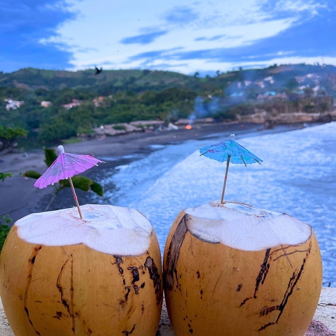 Dos bebidas de coco con sombrillitas en una playa con vista al océano y colinas al fondo.