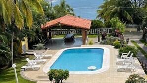 Une piscine avec un pavillon à toit rouge, entourée de palmiers et offrant une vue sur l’océan.