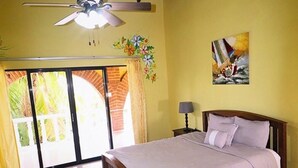 Une chambre à coucher avec un lit, un ventilateur de plafond, une table de chevet, une lampe, un tableau et une vue sur l’extérieur.