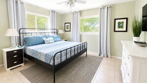 Ein Schlafzimmer mit Bett, Nachttisch, Schminktisch, Deckenventilator und Fenstern mit Vorhängen.