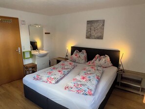 A bedroom with a bed, a bedside table, a nightstand, a mirror, a sink, and a door.
