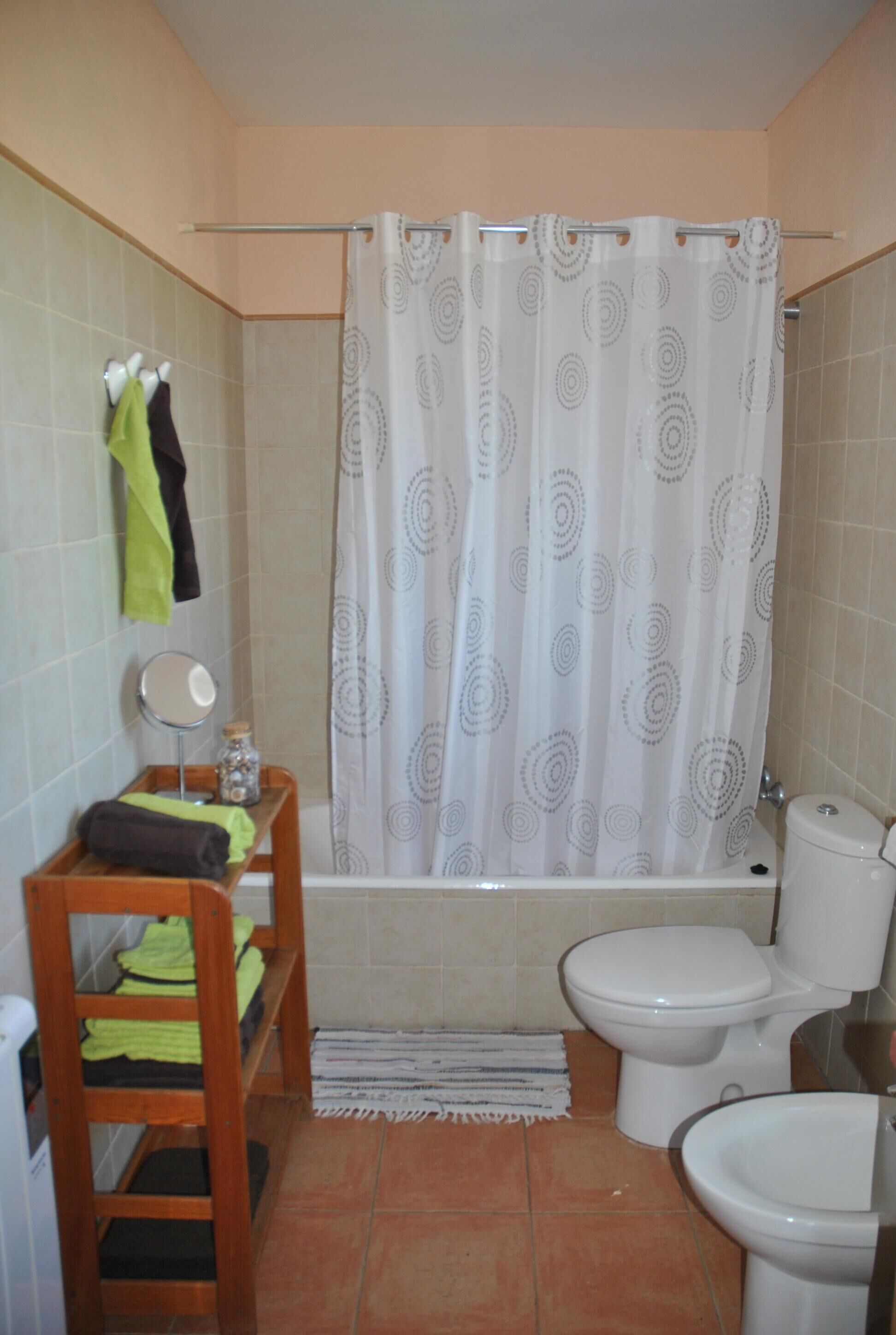 A bathroom with a bathtub, toilet, and sink. There is a shower curtain, a towel rack, and a small table with a mirror and a jar.