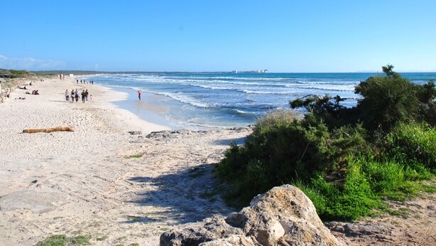 Uma praia com pessoas caminhando pela margem, um afloramento rochoso e um céu azul claro.