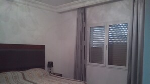 A bedroom with a bed, a nightstand, a window with blinds, and a red pillow. - Very well equipped apartment, 2 bedrooms, living room, guarded parking (EL AOUINA)