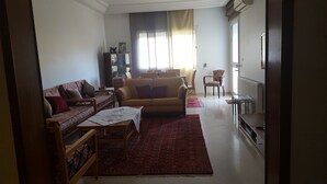 A living room with a sofa, a coffee table, and a TV. There is a window with curtains and a radiator on the wall. - Very well equipped apartment, 2 bedrooms, living room, guarded parking (EL AOUINA)