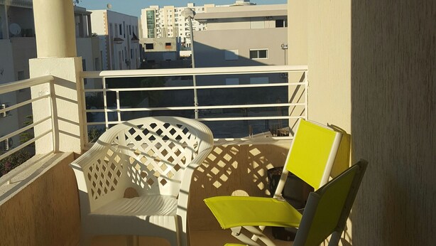A balcony with a white table and two chairs, one yellow and one white, with a view of buildings in the background. - Very well equipped apartment, 2 bedrooms, living room, guarded parking (EL AOUINA)