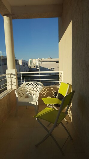 A balcony with a white table and two chairs, one yellow and one white, with a view of buildings in the background. - Very well equipped apartment, 2 bedrooms, living room, guarded parking (EL AOUINA)