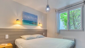 Un dormitorio con cama, lámparas de noche, una mesita de noche, un cuadro de un barco en el agua y una ventana con vista a árboles.