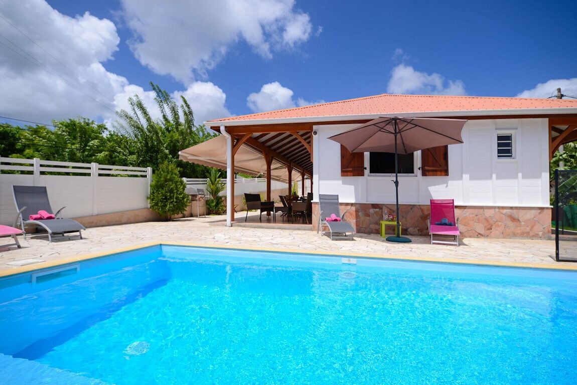 Una piscina con sillas de descanso y sombrillas, un patio cubierto y un muro de piedra.