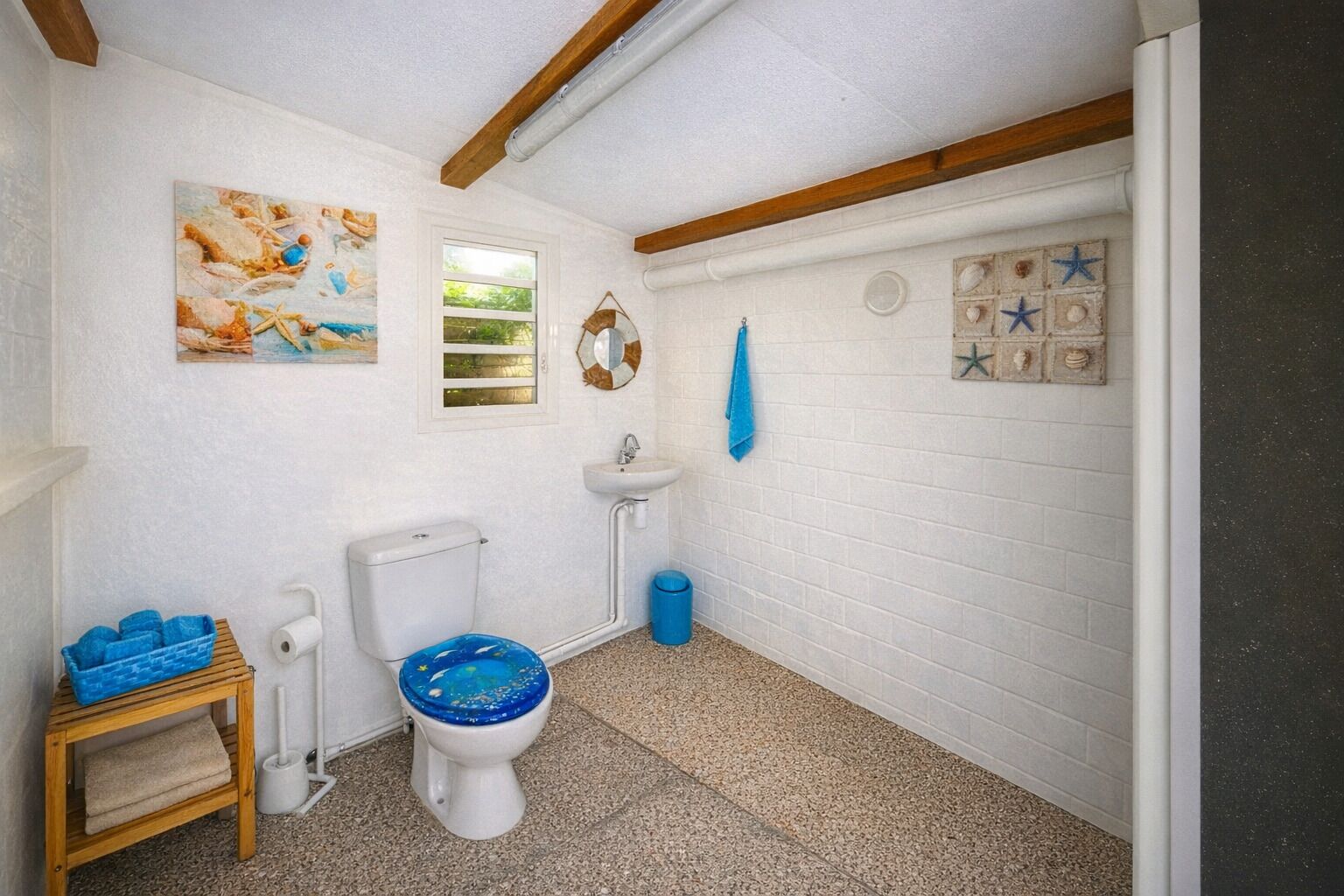 A bathroom with a white toilet, a sink, a blue trash bin, and a wooden shelf with towels.