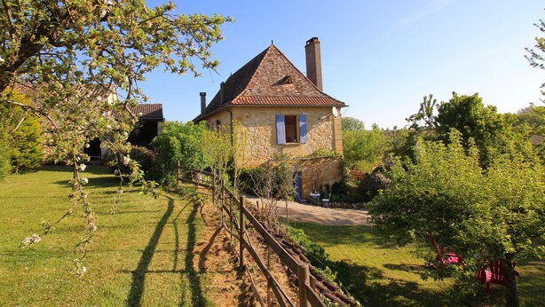 Une maison en pierre avec un toit en tuiles, entourée d’un jardin bien entretenu et d’une clôture en bois.