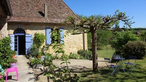 Une maison en pierre avec un toit en tuiles, un jardin agrémenté de plantes en pot et une table avec des chaises à l’extérieur.