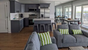 A modern kitchen with grey cabinets, stainless steel appliances, and a central island. There is a dining area with a table and chairs, and a balcony with a view. - 2 BR Bayfront Condo with Gorgeous Sunsets, in Ocean City NJ (Ocean City)
