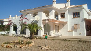 Une maison à deux étages, à l’extérieur blanc, dotée de plusieurs balcons et d’un petit jardin agrémenté de rochers et d’un palmier.