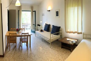 A bedroom with a bed, bedside table, and a small desk with a chair. There is a window with curtains, a door, and a balcony visible.