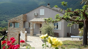 A stone house with a stone staircase leading to a covered porch, surrounded by lush greenery and mountains in the background. - LARGE VILLA - 4 **** - HEATED SWIMMING POOL, 180° SEA VIEW - AIR CONDITIONED 
 (Sisco)