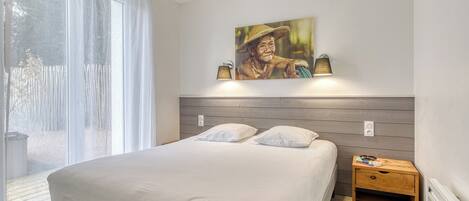 Une chambre à coucher comprenant un lit, une table de chevet, une lampe fixée au mur, un tableau et une fenêtre avec des rideaux légers.
