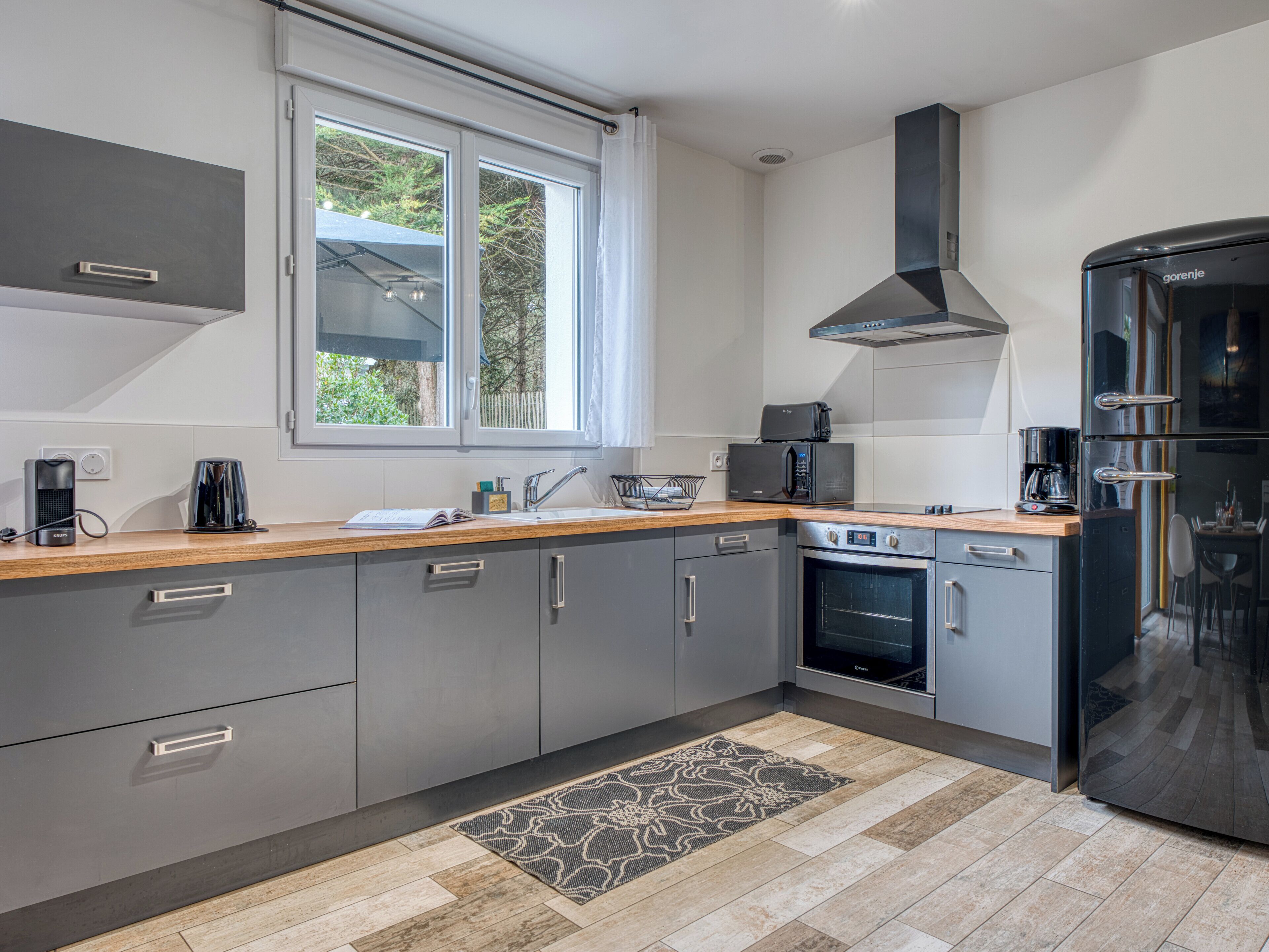 A modern kitchen with grey cabinets, a built-in oven, a black refrigerator, and a window with a view of greenery.