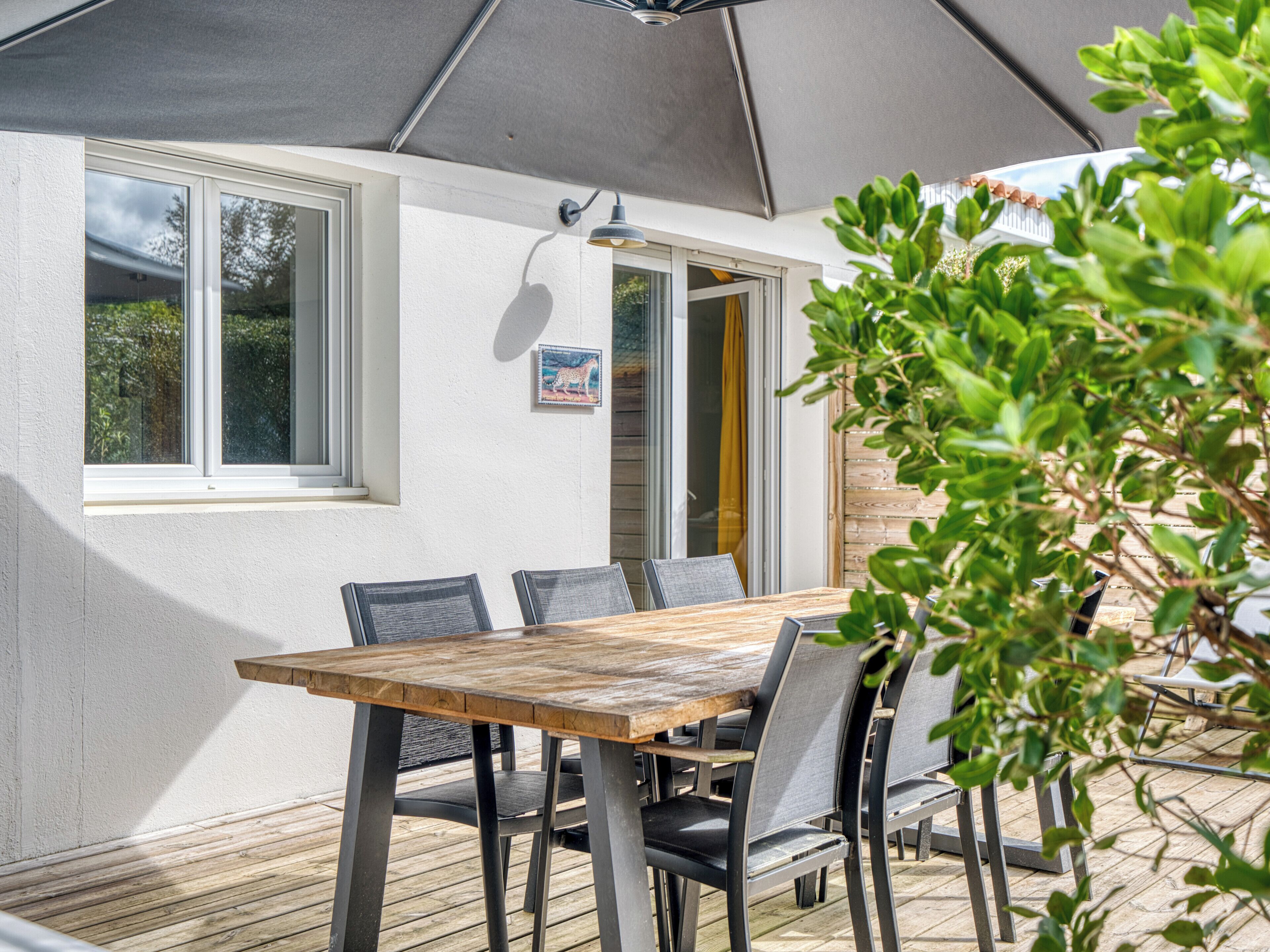 An outdoor patio with a wooden table and chairs, a large umbrella, and a window with a view of greenery.