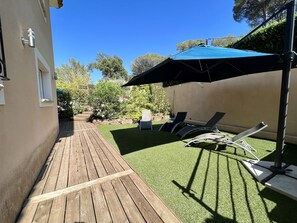 Une terrasse en bois, un parasol bleu et deux chaises longues dans un jardin.