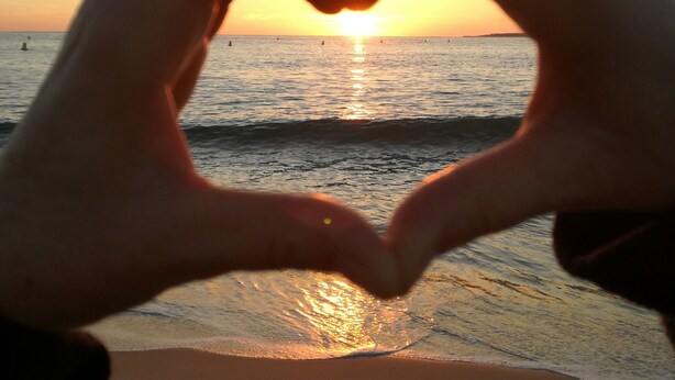 Hands forming a heart shape in front of a sunset over the ocean.