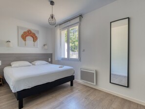 A bedroom with a bed, a window, a wall-mounted mirror, and a ceiling light fixture. - Villa Russie - Domaine Le Sherwood (Notre-Dame-De-Monts)
