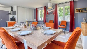 A dining area with a wooden table set for six, orange chairs, a kitchen in the background, and a large window with curtains.