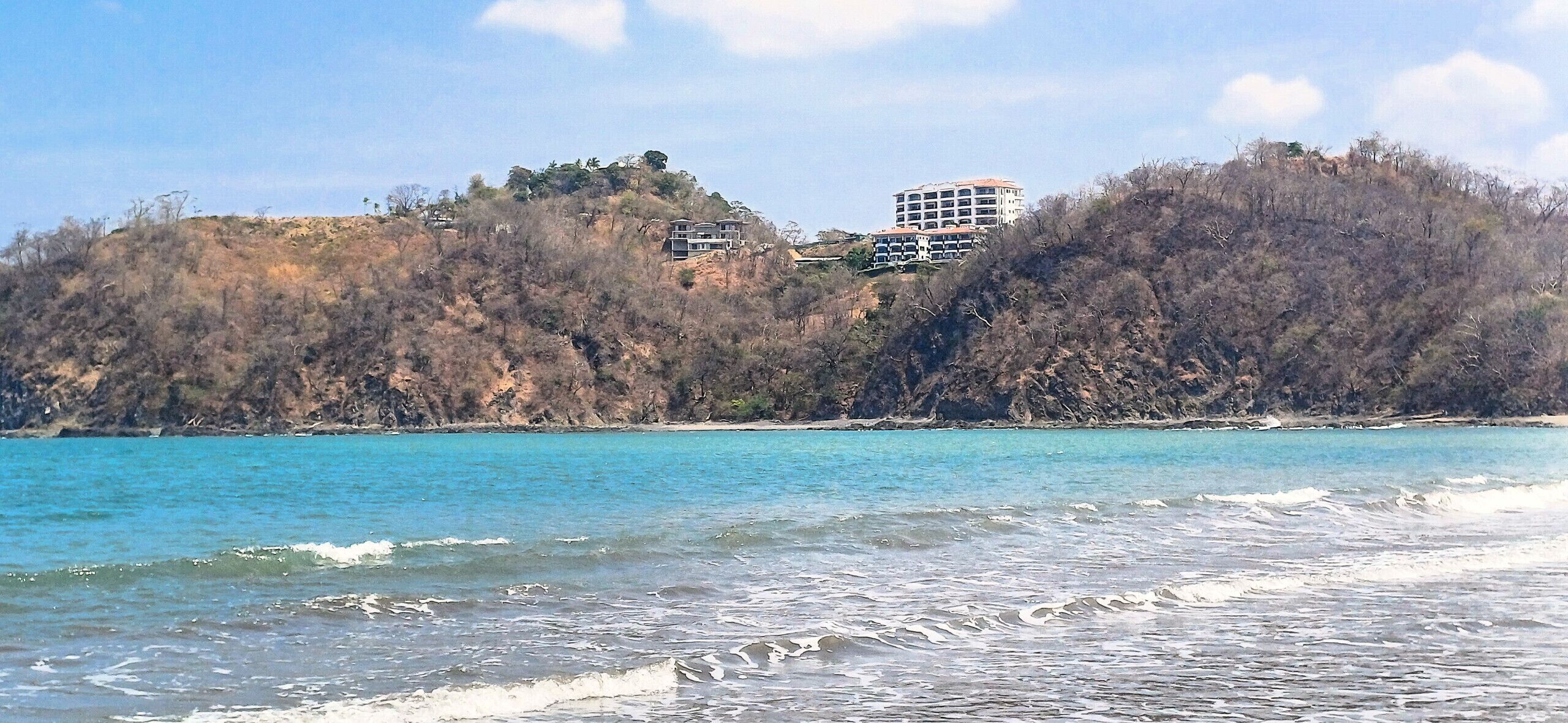 A coastal view with a building on a hill, overlooking a beach.