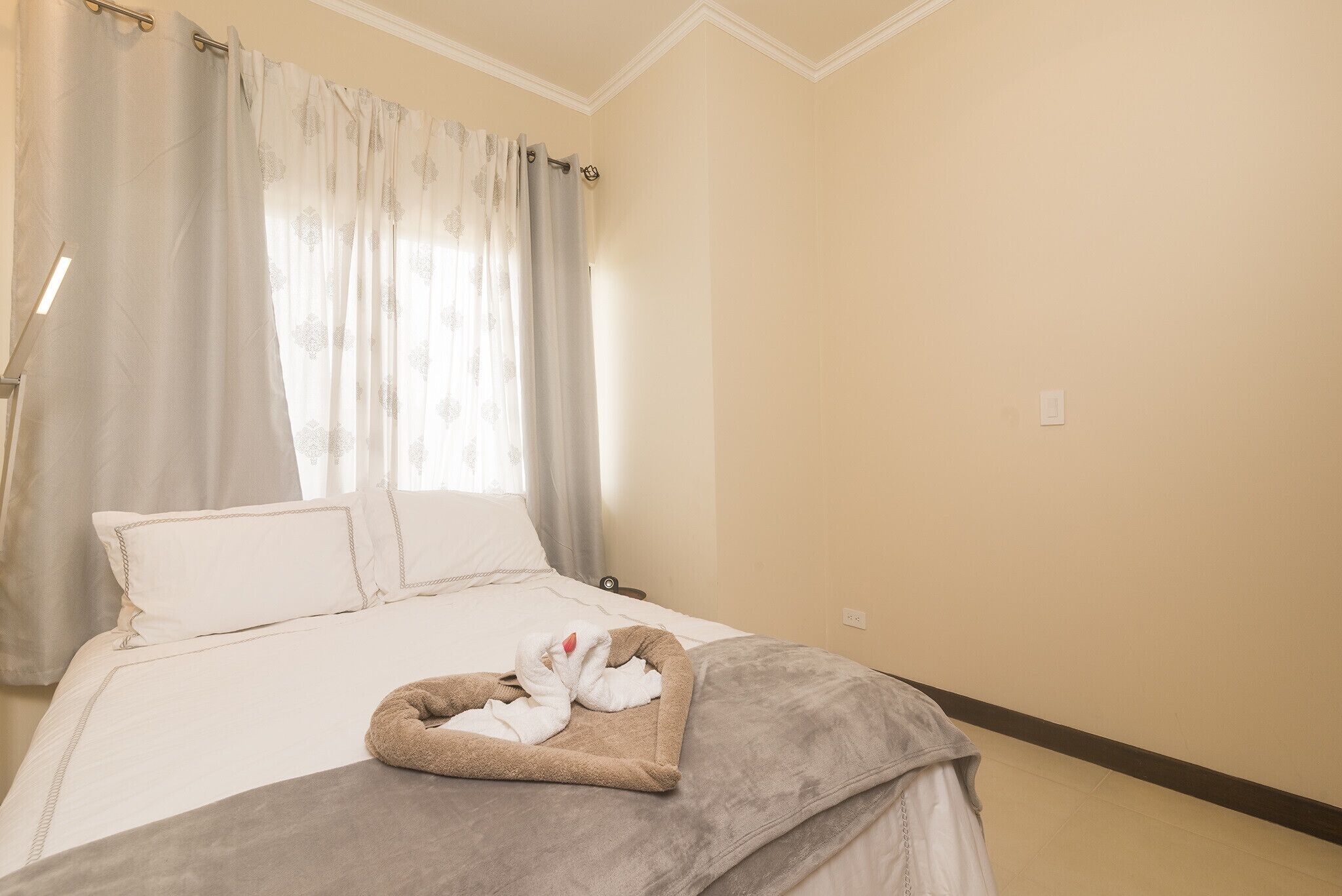 A neatly made bed with a heart-shaped towel arrangement, a bedside table, and a window with curtains.