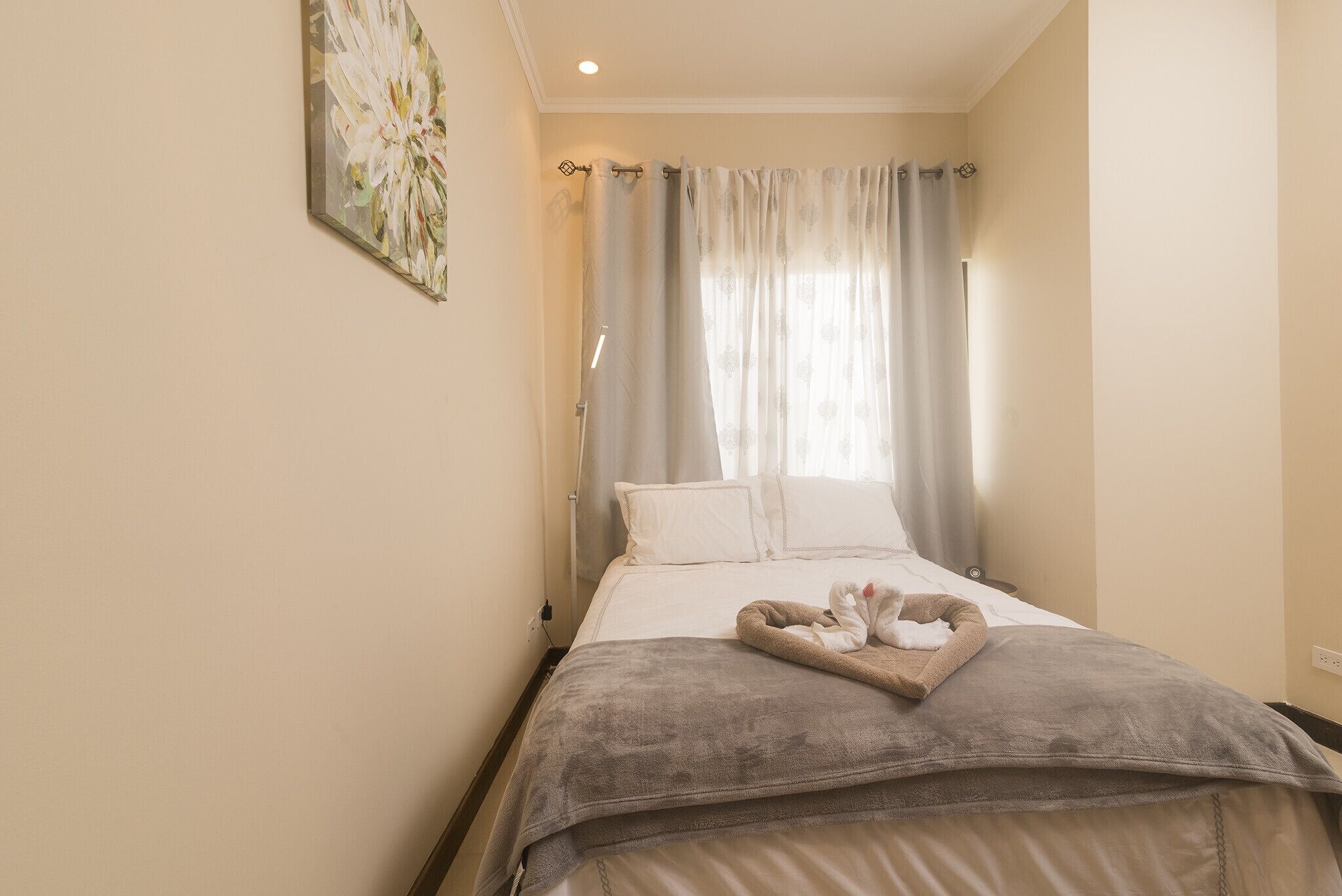 Una cama bien hecha con una manta gris y una toalla, un cuadro en la pared y una ventana con cortinas.