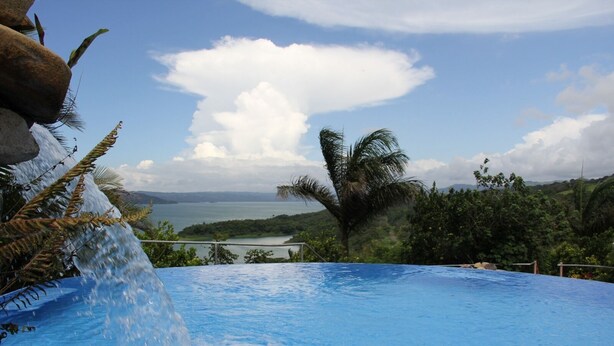 A swimming pool with a waterfall feature, surrounded by lush greenery and a view of a body of water and distant mountains. - 2B/1B CORNER CONDO SUITE with VIEWS (Tilaran)
