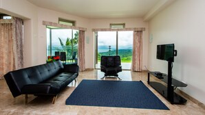 A spacious living room with a black leather sofa, a blue exercise mat, a black office chair, and a flat-screen TV. Large windows offer a view of the outdoors. - 2B/1B CORNER CONDO SUITE with VIEWS (Tilaran)