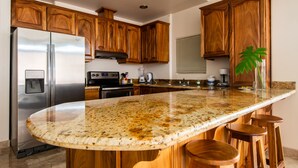 A kitchen with wooden cabinets, a stainless steel refrigerator, a granite countertop, and wooden stools. - 2B/1B CORNER CONDO SUITE with VIEWS (Tilaran)