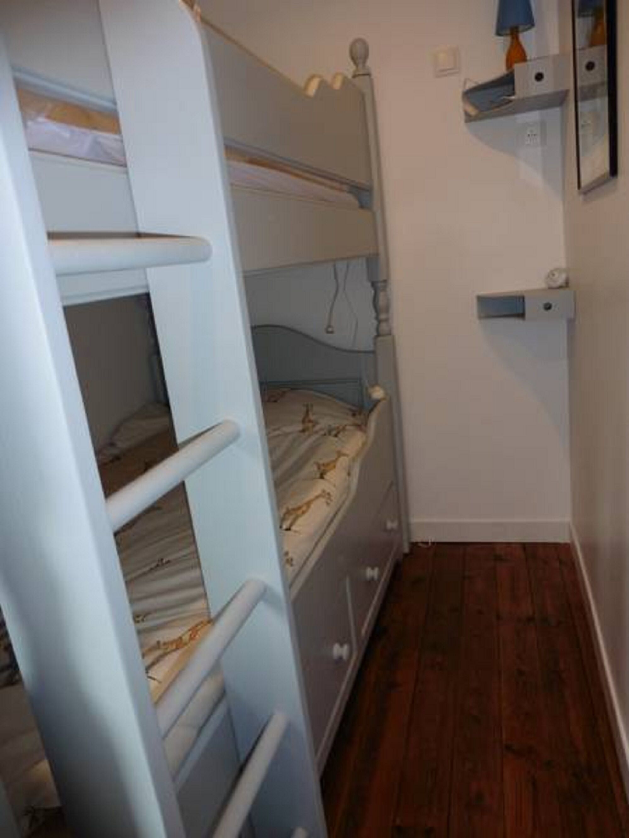 Habitación con literas, suelo de madera, una pequeña estantería con lámpara y un espejo en la pared.