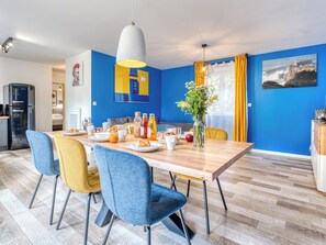 A modern dining area with a wooden table, blue and yellow chairs, and a kitchen in the background.