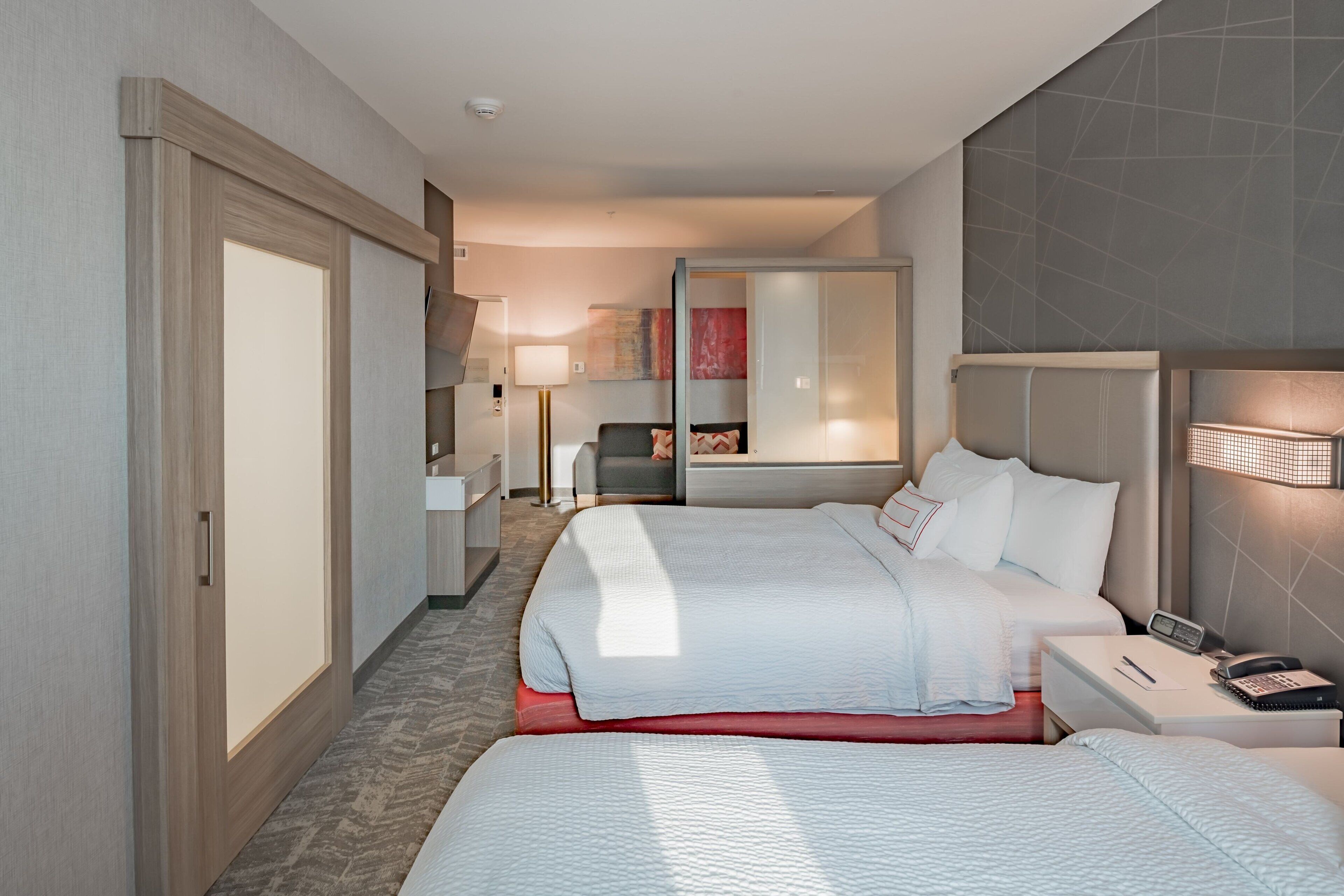 A hotel room with two beds, a desk, a chair, a mirror, and a bathroom.