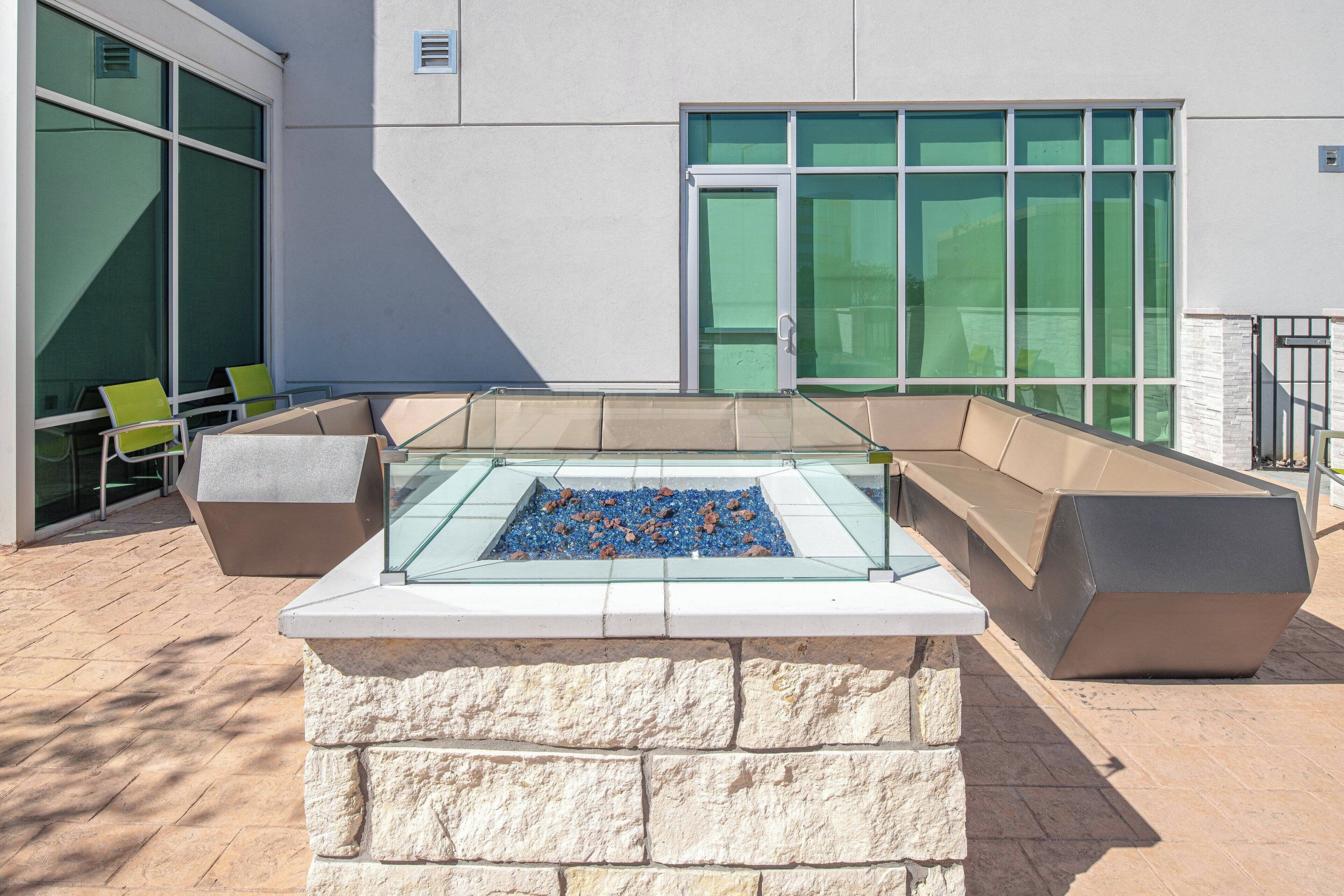 An outdoor seating area with a fire pit, stone walls, and a building with large windows in the background.