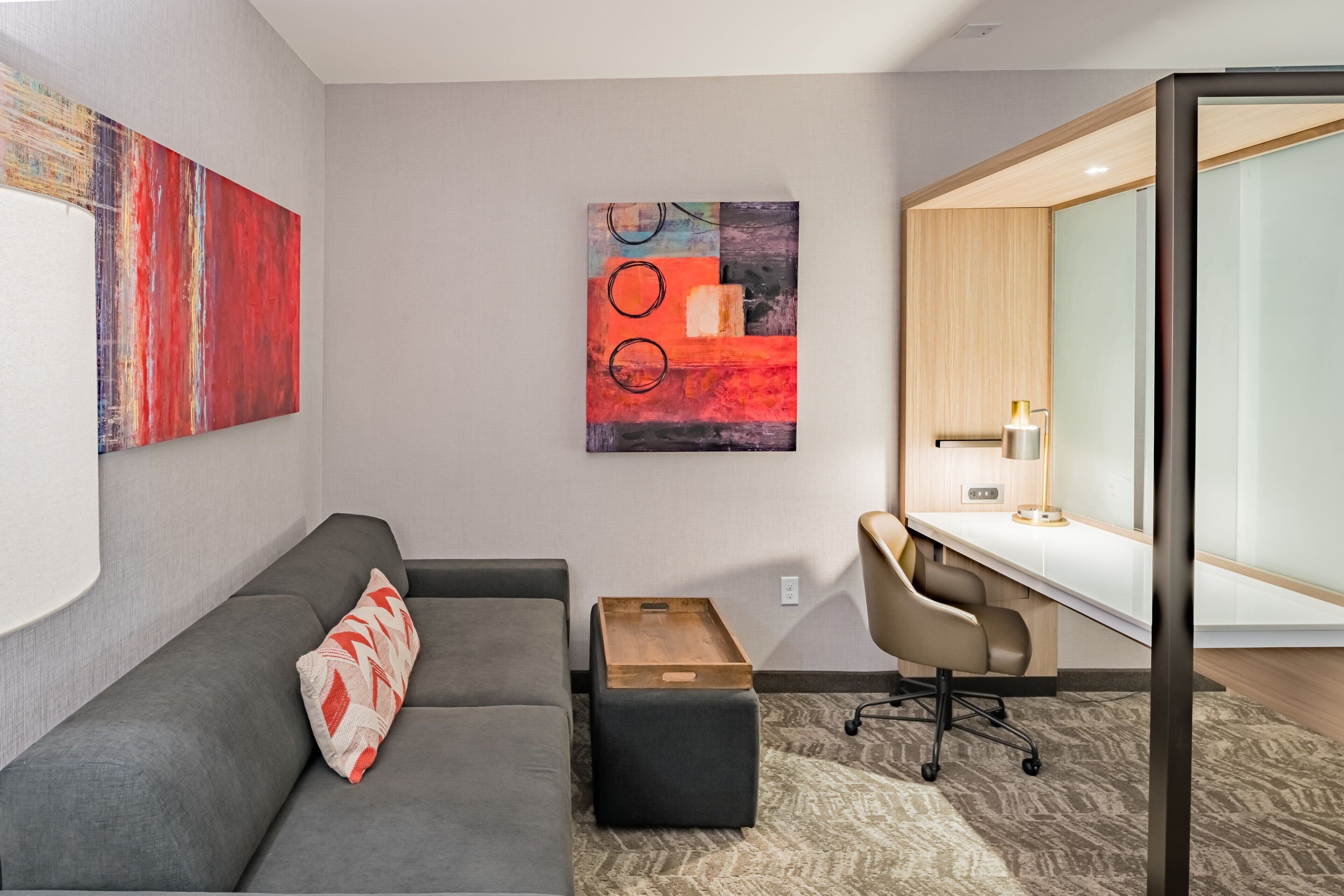 A modern room with a grey sofa, a wooden side table, a desk with a chair, and abstract art on the walls.