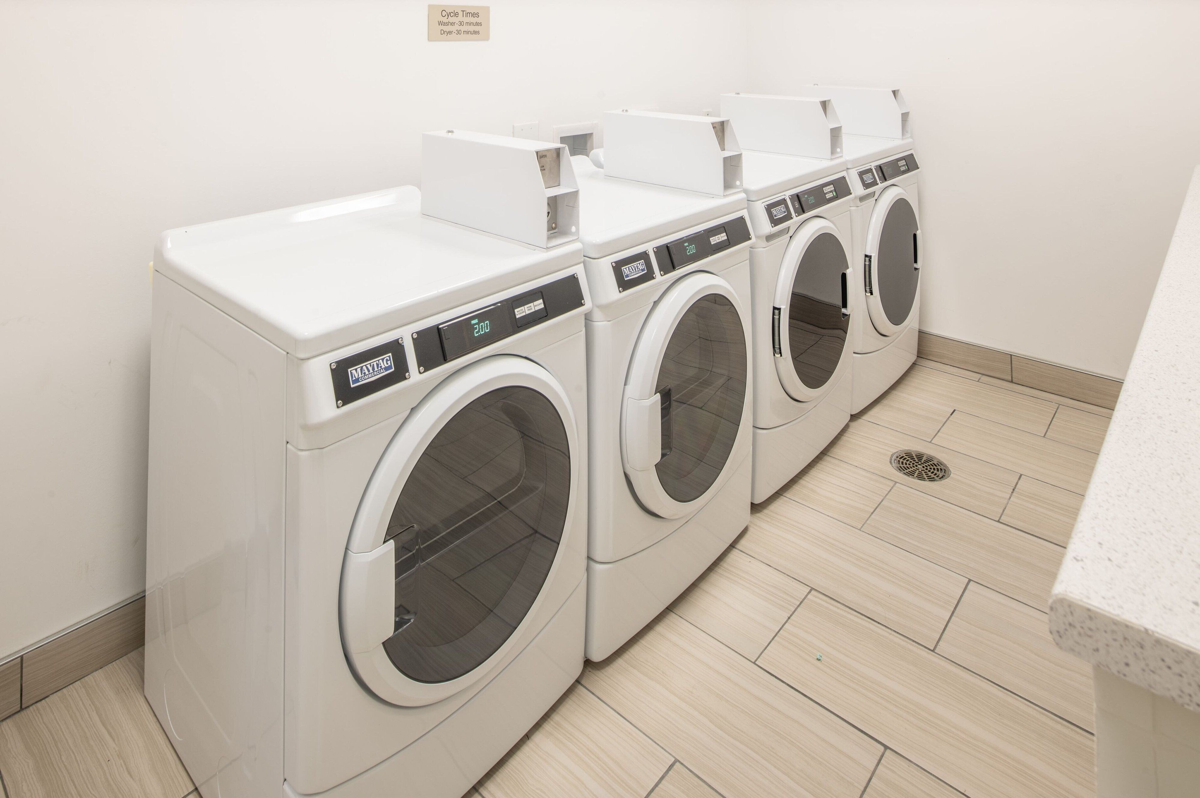 A row of white front-loading washing machines in a laundry room with a tiled floor and a white wall.