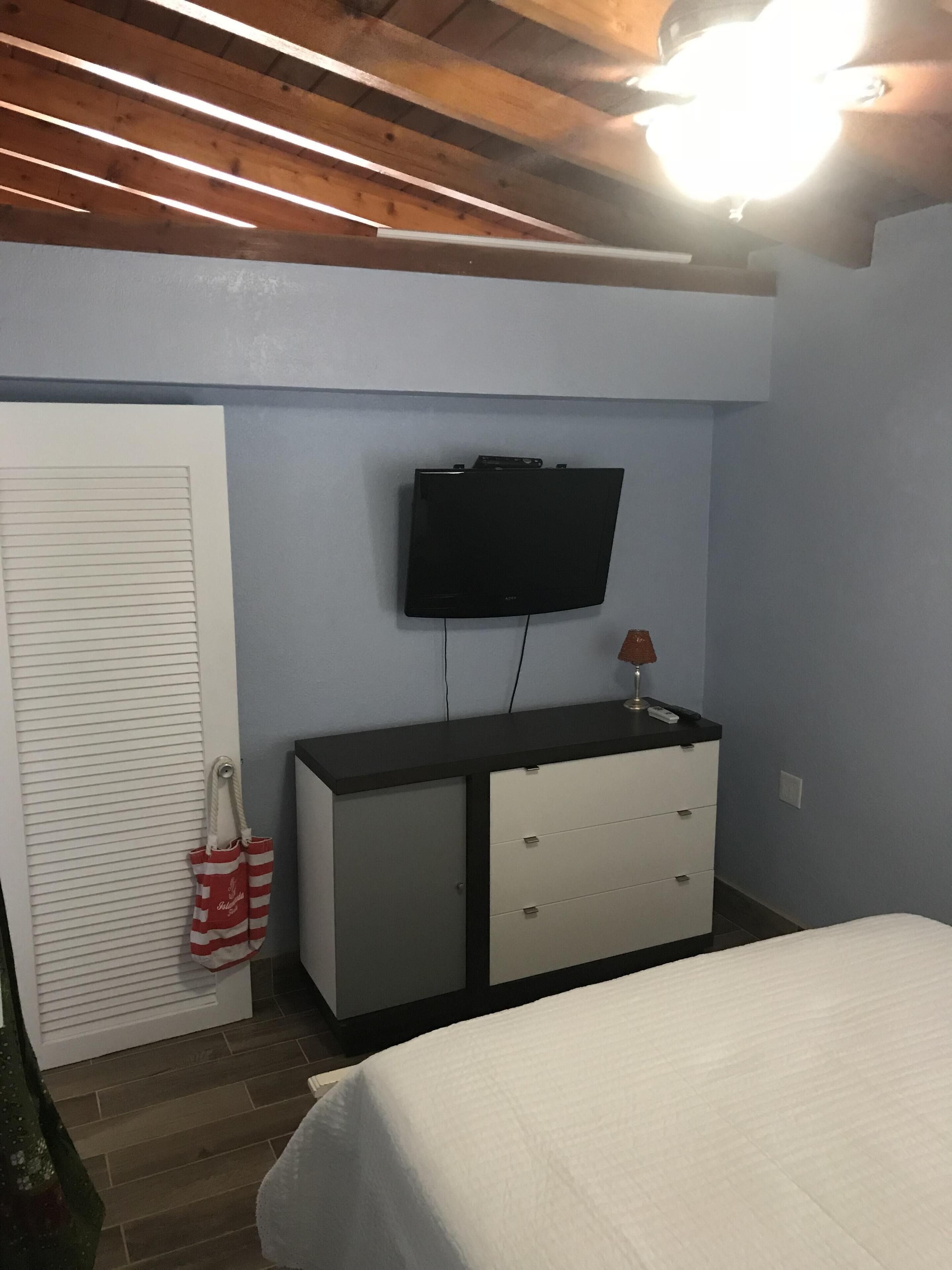 A bedroom with a bed, a TV mounted on a wall, a dresser, and a door with a striped bag hanging on it.