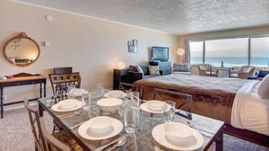 Una camera d'albergo con un letto grande, un'area pranzo con tavolo apparecchiato per quattro persone, una televisione e vista sull'oceano.